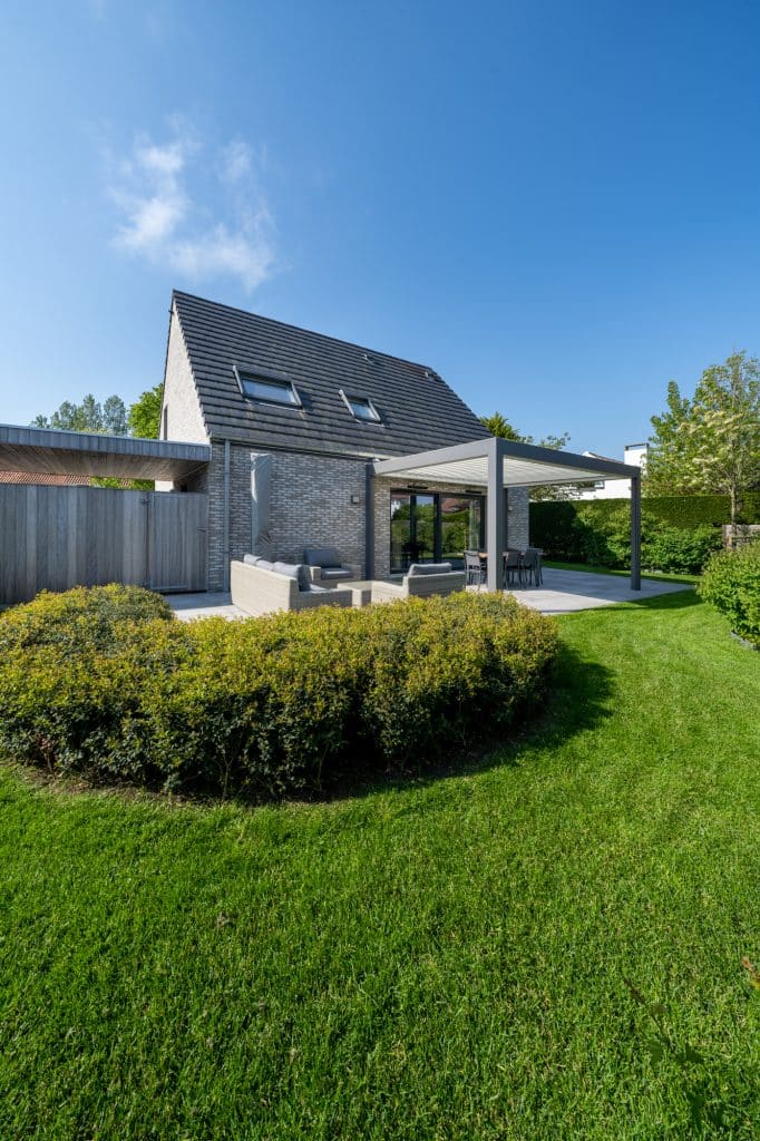 Modern nieuwbouw huis in Koksijde met een opvallend asymmetrisch dak, een terras, overkapping en een grote, strakke groene tuin onder een blauwe lucht