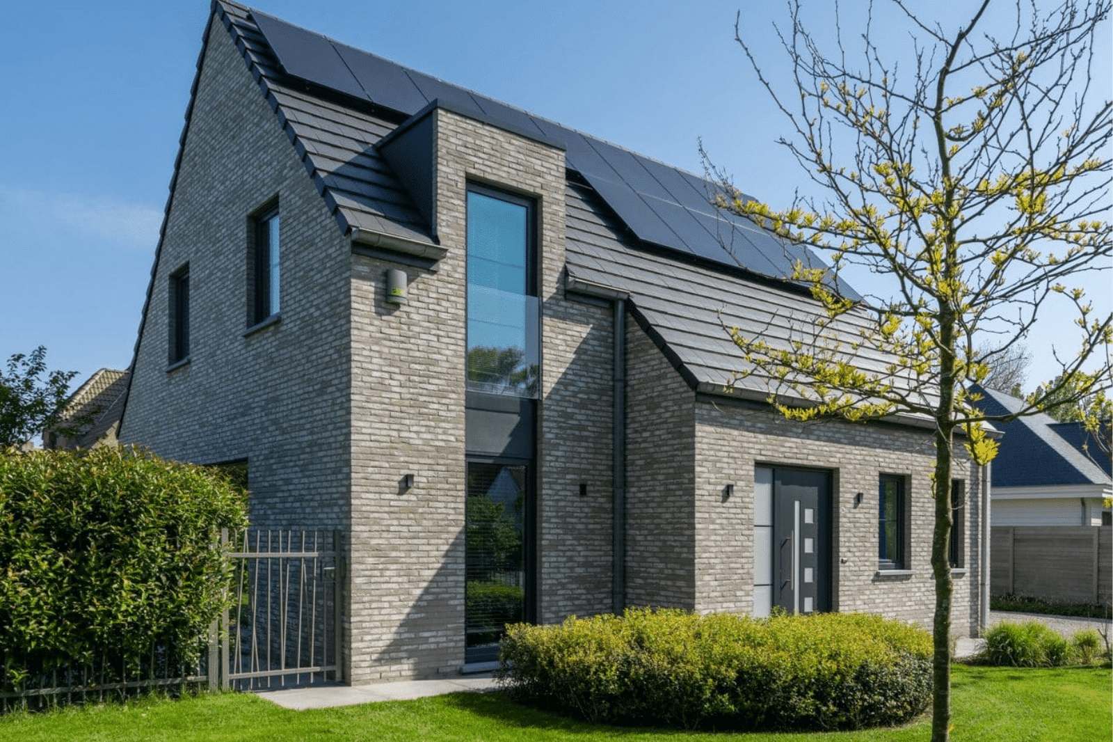 Eigentijdse sleutel-op-de-deur woning in lichte gevelsteen met zwart hellend dak, zonnepanelen en een hoog raam naast de voordeur.
