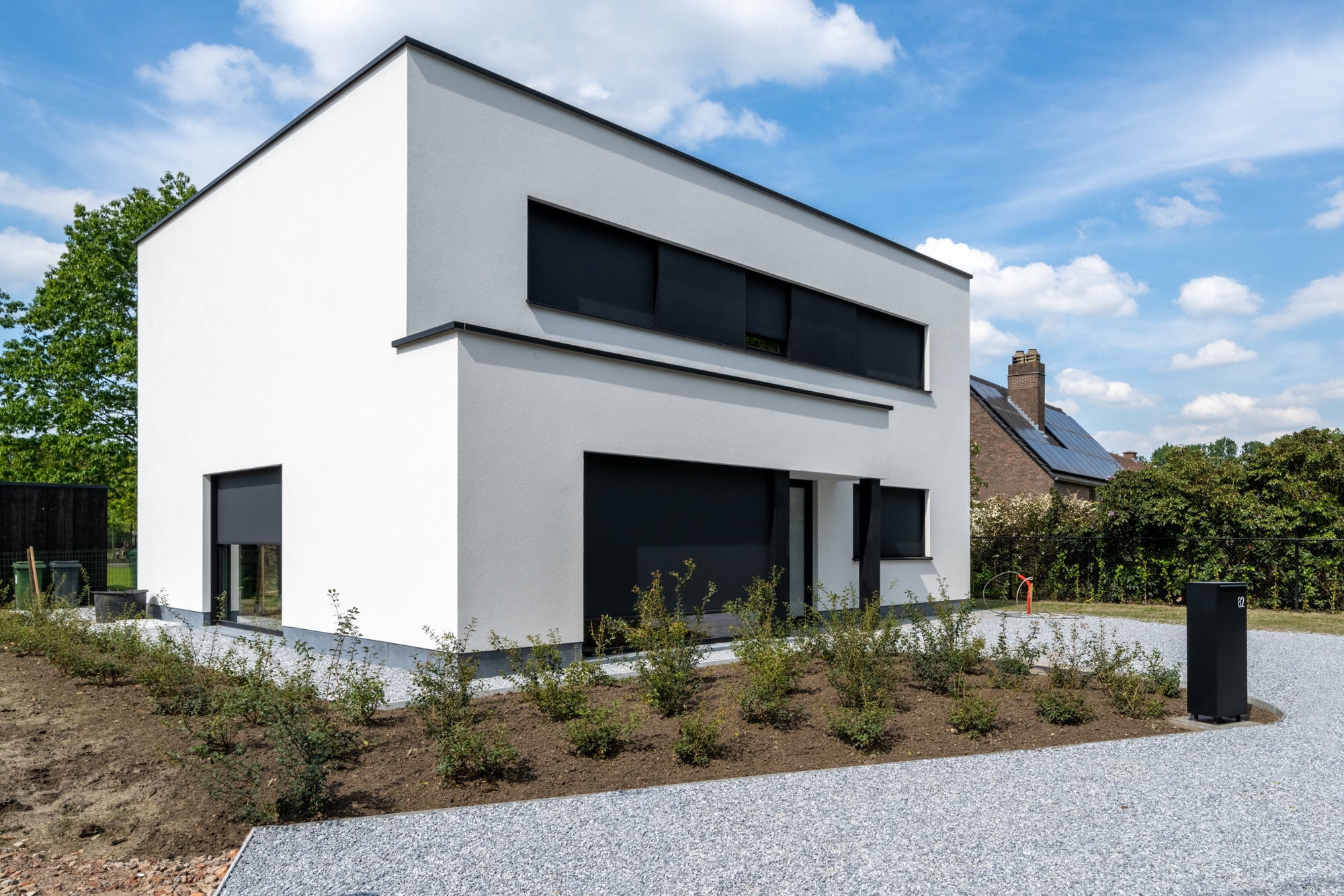 Kubusvormige nieuwbouwwoning in het wit met plat dak en zwarte zonwerende screens, omgeven door een pas aangelegde voortuin.