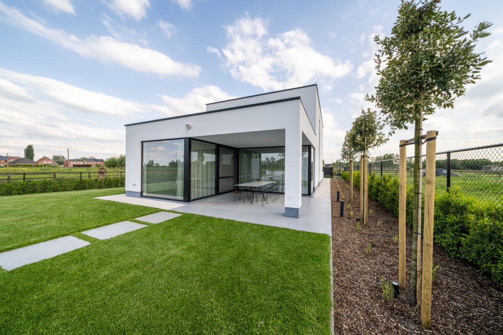 Moderne witte nieuwbouwwoning met plat dak en overdekt terras met eettafel, uitkijkend over een ruime, groene tuin.