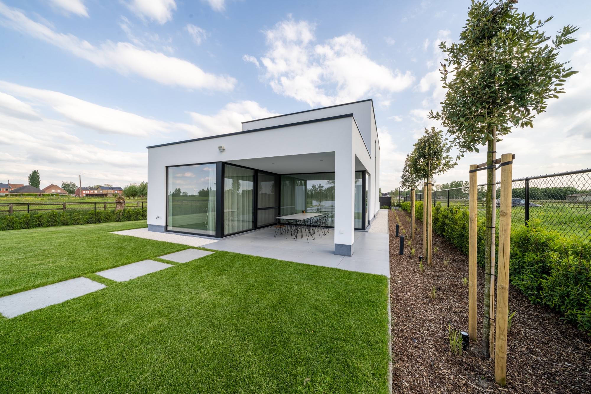 Moderne witte nieuwbouwwoning met plat dak en overdekt terras met eettafel, uitkijkend over een ruime, groene tuin.